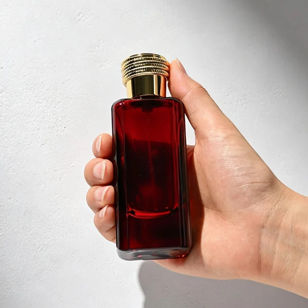 two tall rectangular, thick-bottomed glass perfume bottles are shown standing side by side. One bottle is made of vivid semi-translucent red glass and the other is vibrant semi-translucent green glass. Each bottle features subtly debossed vertical panels along its sides and is topped with a cylindrical cap encircled by a textured, coiling snake pattern—presented in polished gold on the red bottle and polished silver on the green bottle.
