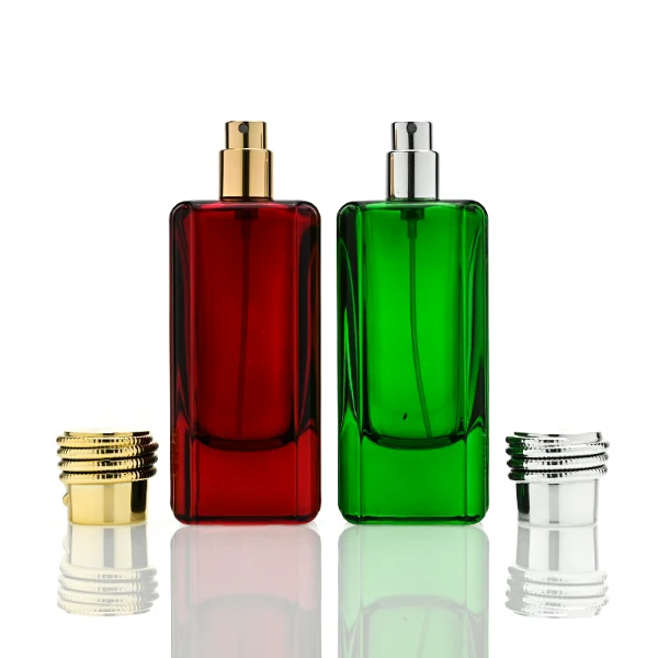two tall rectangular, thick-bottomed glass perfume bottles are shown standing side by side. One bottle is made of vivid semi-translucent red glass and the other is vibrant semi-translucent green glass. Each bottle features subtly debossed vertical panels along its sides and is topped with a cylindrical cap encircled by a textured, coiling snake pattern—presented in polished gold on the red bottle and polished silver on the green bottle.