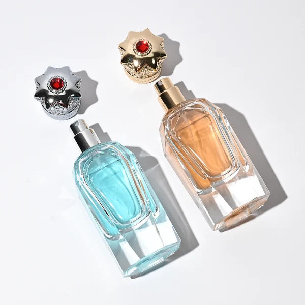 Three clear glass perfume bottles with faceted, angular bodies and heavy, thick bases are shown containing amber, light blue, and olive green liquids. Each bottle is topped with an ornate, highly detailed Zamac crown-motif cap featuring intricate filigree metalwork and a small red pointed gem accent at the peak, presented in polished gold, bright silver, and dark gunmetal finishes.
