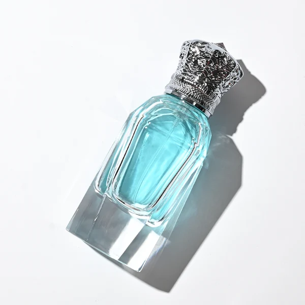 Three clear glass perfume bottles with faceted, angular bodies and heavy, thick bases are shown containing amber, light blue, and olive green liquids. Each bottle is topped with an ornate, highly detailed Zamac crown-motif cap featuring intricate filigree metalwork and a small red pointed gem accent at the peak, presented in polished gold, bright silver, and dark gunmetal finishes.