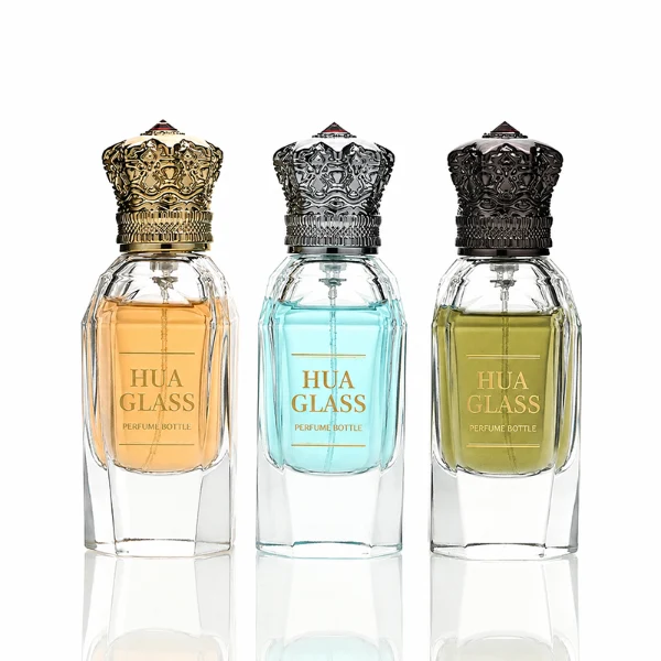 Three clear glass perfume bottles with faceted, angular bodies and heavy, thick bases are shown containing amber, light blue, and olive green liquids. Each bottle is topped with an ornate, highly detailed Zamac crown-motif cap featuring intricate filigree metalwork and a small red pointed gem accent at the peak, presented in polished gold, bright silver, and dark gunmetal finishes.
