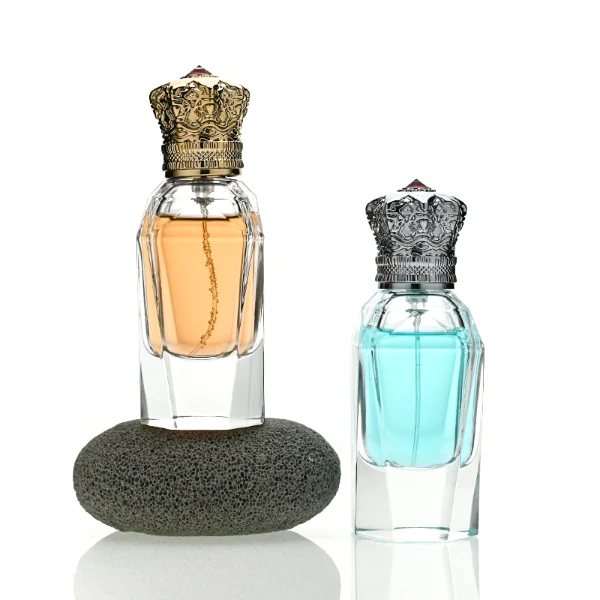 Three clear glass perfume bottles with faceted, angular bodies and heavy, thick bases are shown containing amber, light blue, and olive green liquids. Each bottle is topped with an ornate, highly detailed Zamac crown-motif cap featuring intricate filigree metalwork and a small red pointed gem accent at the peak, presented in polished gold, bright silver, and dark gunmetal finishes.