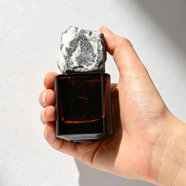 Two amber-brown cylindrical glass perfume bottles are shown in different sizes. The smaller bottle features a rugged charcoal gray natural stone cap, while the larger bottle features a craggy, tan and golden-brown natural rock cap.
