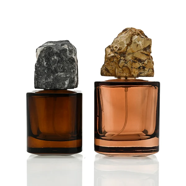 Two amber-brown cylindrical glass perfume bottles are shown in different sizes. The smaller bottle features a rugged charcoal gray natural stone cap, while the larger bottle features a craggy, tan and golden-brown natural rock cap.