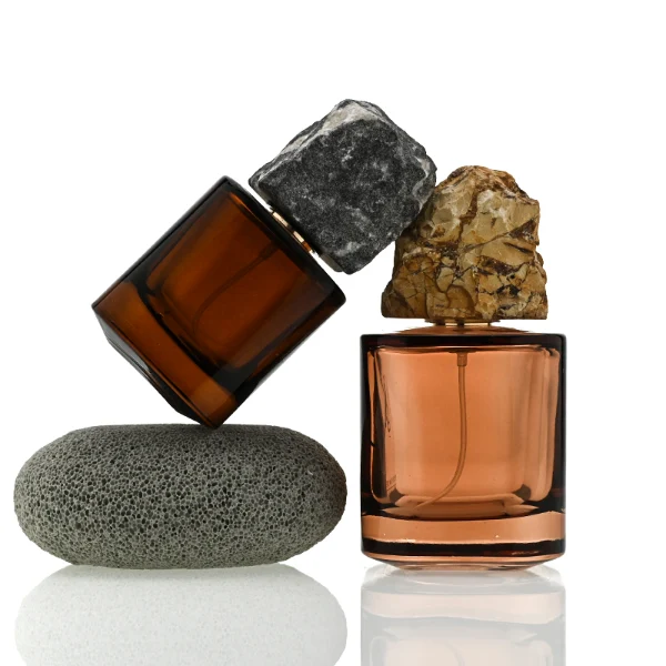 Two amber-brown cylindrical glass perfume bottles are shown in different sizes. The smaller bottle features a rugged charcoal gray natural stone cap, while the larger bottle features a craggy, tan and golden-brown natural rock cap.