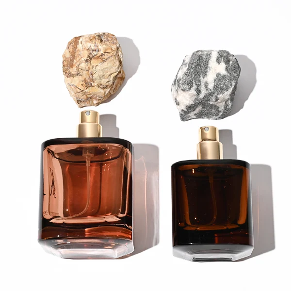 Two amber-brown cylindrical glass perfume bottles are shown in different sizes. The smaller bottle features a rugged charcoal gray natural stone cap, while the larger bottle features a craggy, tan and golden-brown natural rock cap.