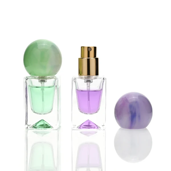 Three small, short rectangular clear glass perfume bottles with thick bases stand side by side, containing light purple, amber, and mint green liquids. Each bottle is topped with a large, glossy spherical resin cap featuring marble swirl patterns in matching shades of lavender, dark tortoiseshell brown, and pale sage green.