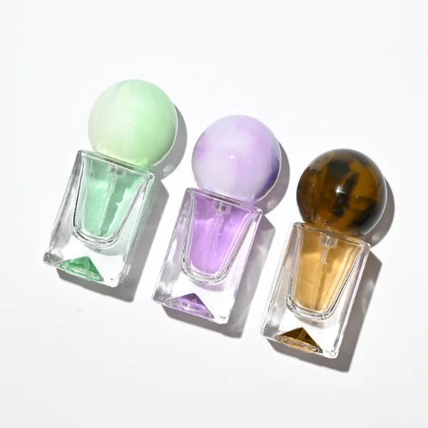 Three small, short rectangular clear glass perfume bottles with thick bases stand side by side, containing light purple, amber, and mint green liquids. Each bottle is topped with a large, glossy spherical resin cap featuring marble swirl patterns in matching shades of lavender, dark tortoiseshell brown, and pale sage green.