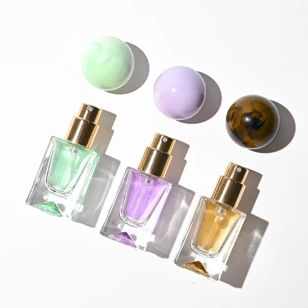 Three small, short rectangular clear glass perfume bottles with thick bases stand side by side, containing light purple, amber, and mint green liquids. Each bottle is topped with a large, glossy spherical resin cap featuring marble swirl patterns in matching shades of lavender, dark tortoiseshell brown, and pale sage green.