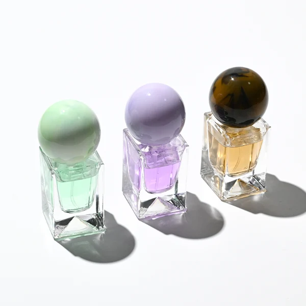 Three small, short rectangular clear glass perfume bottles with thick bases stand side by side, containing light purple, amber, and mint green liquids. Each bottle is topped with a large, glossy spherical resin cap featuring marble swirl patterns in matching shades of lavender, dark tortoiseshell brown, and pale sage green.