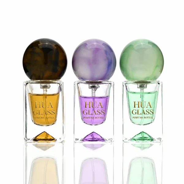 Three small, short rectangular clear glass perfume bottles with thick bases stand side by side, containing light purple, amber, and mint green liquids. Each bottle is topped with a large, glossy spherical resin cap featuring marble swirl patterns in matching shades of lavender, dark tortoiseshell brown, and pale sage green.