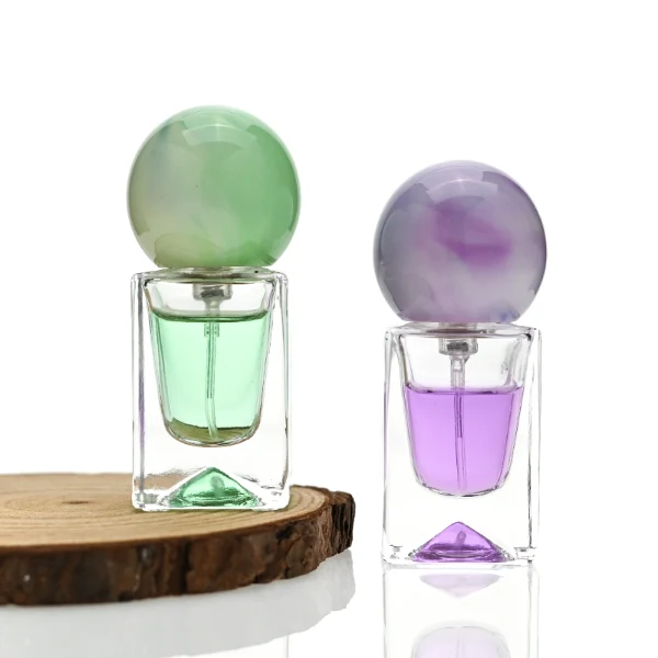 Three small, short rectangular clear glass perfume bottles with thick bases stand side by side, containing light purple, amber, and mint green liquids. Each bottle is topped with a large, glossy spherical resin cap featuring marble swirl patterns in matching shades of lavender, dark tortoiseshell brown, and pale sage green.