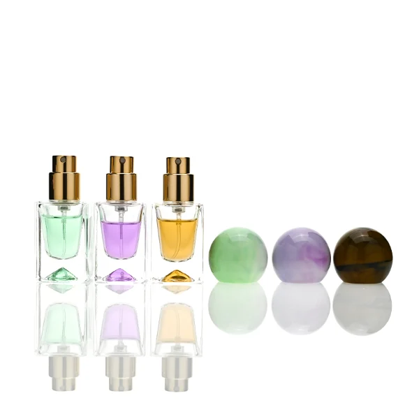 Three small, short rectangular clear glass perfume bottles with thick bases stand side by side, containing light purple, amber, and mint green liquids. Each bottle is topped with a large, glossy spherical resin cap featuring marble swirl patterns in matching shades of lavender, dark tortoiseshell brown, and pale sage green.