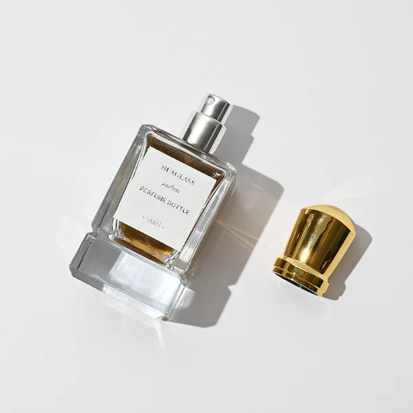 HUA0058 perfume bottle, heavy-base rectangular cuboid glass bottle with clear finish, featuring a paper label, a matching neck, and a tall sculptural cylindrical plastic cap with a sharply angled sloping top in a smooth high-gloss shiny gold finish, elegant modern fragrance packaging design available in 30ml, 50ml, and 100ml