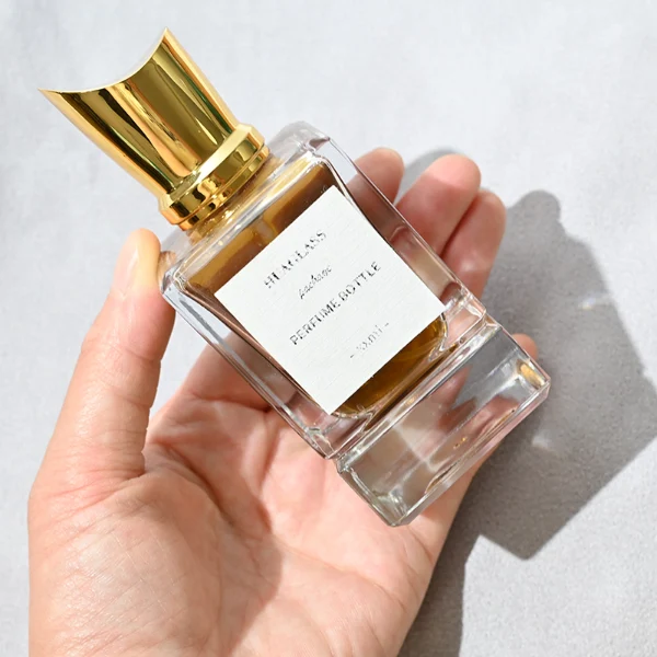 HUA0058 perfume bottle, heavy-base rectangular cuboid glass bottle with clear finish, featuring a paper label, a matching neck, and a tall sculptural cylindrical plastic cap with a sharply angled sloping top in a smooth high-gloss shiny gold finish, elegant modern fragrance packaging design available in 30ml, 50ml, and 100ml