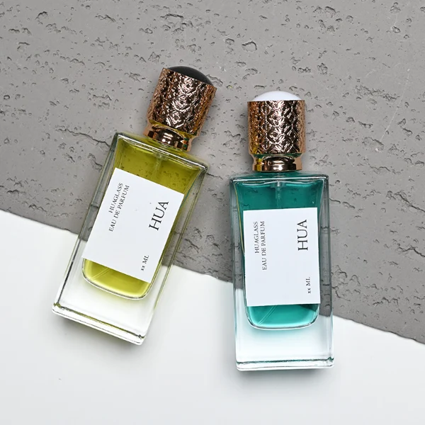 Two thick-bottomed rectangular clear glass perfume bottles stand side by side, containing bright yellow and teal liquids. Both bottles feature a square white label with the text "HUA" and are topped with cylindrical golden caps that showcase an intricate, textured diamond lattice pattern.