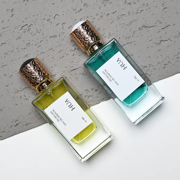 Two thick-bottomed rectangular clear glass perfume bottles stand side by side, containing bright yellow and teal liquids. Both bottles feature a square white label with the text "HUA" and are topped with cylindrical golden caps that showcase an intricate, textured diamond lattice pattern.