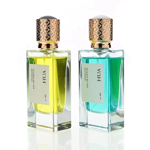 Two thick-bottomed rectangular clear glass perfume bottles stand side by side, containing bright yellow and teal liquids. Both bottles feature a square white label with the text "HUA" and are topped with cylindrical golden caps that showcase an intricate, textured diamond lattice pattern.