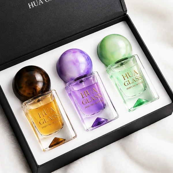 Three small, short rectangular clear glass perfume bottles with thick bases stand side by side, containing light purple, amber, and mint green liquids. Each bottle is topped with a large, glossy spherical resin cap featuring marble swirl patterns in matching shades of lavender, dark tortoiseshell brown, and pale sage green.